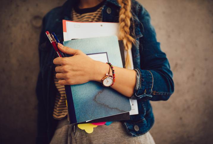 Teenage girl holding books, notebooks and pencils standing against wall