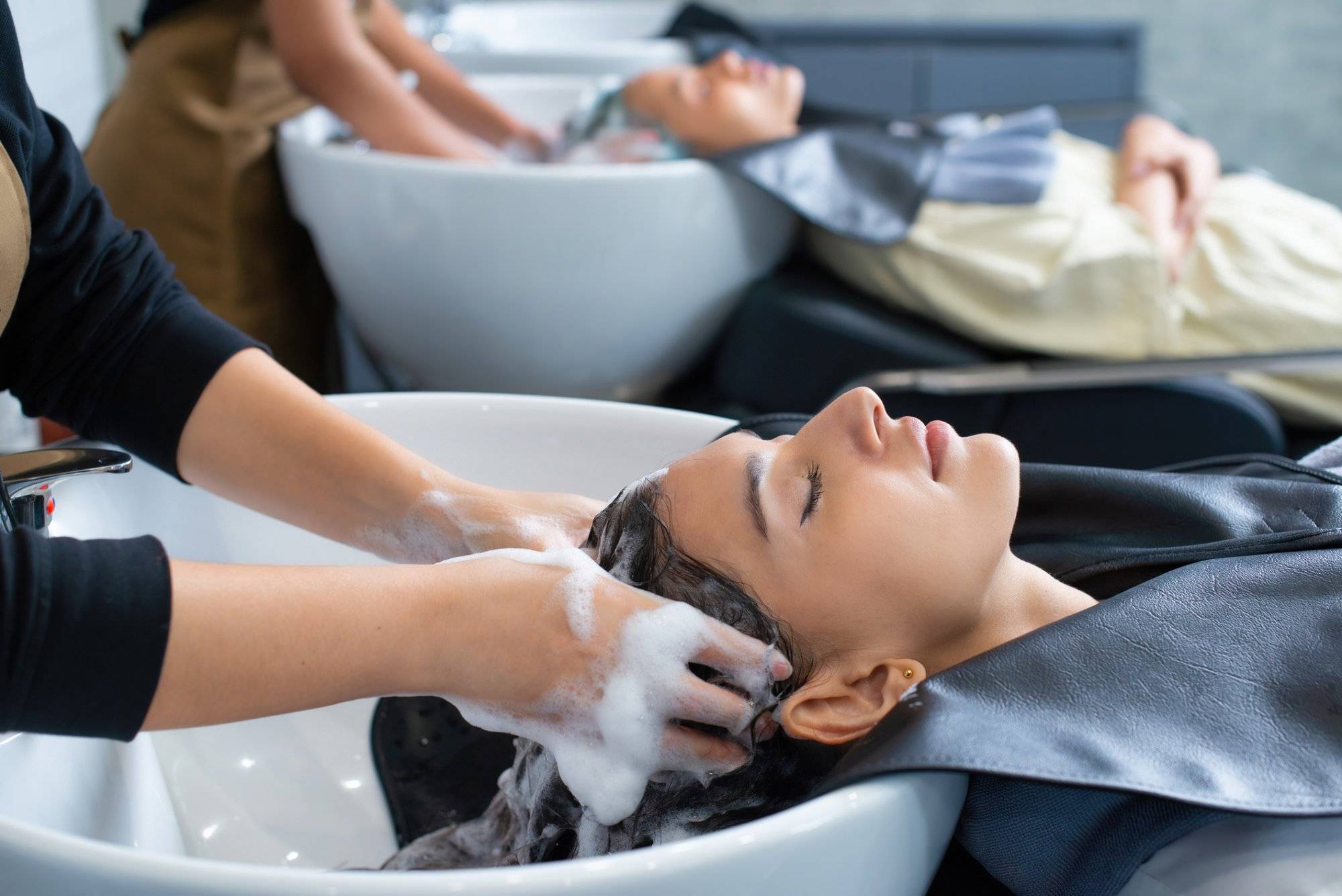 Young woman in hairdresser salon during hair wash after haircut. Woman applying shampoo and massaging hair of a customer. Female having her hair washed in a hairdressing salon.